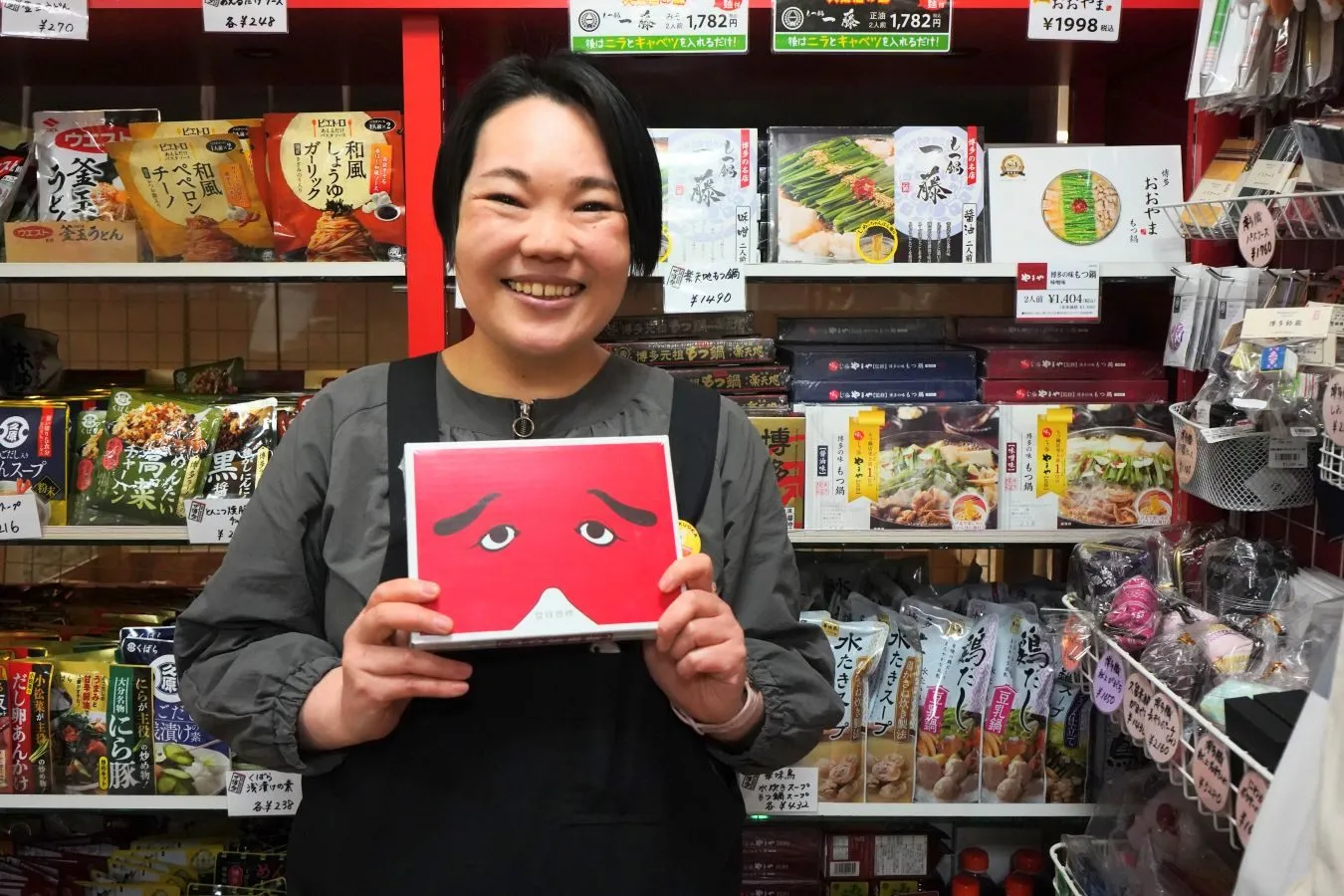 ▲“博多仁和加（はかたにわか）”の半面を形どったユーモアあふれる「二〇加煎餅（にわかせんぺい）」（東雲堂）を手にする藤嶋さん